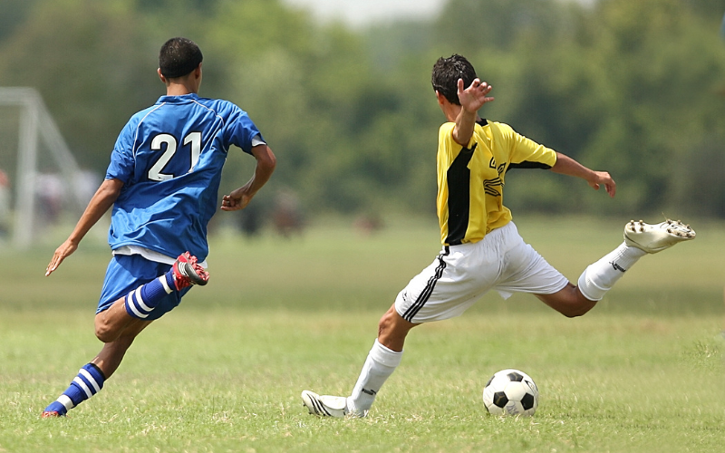 Two men are playing soccer on a field, one is wearing a blue uniform and the other is wearing a yellow uniform, kicking a ball