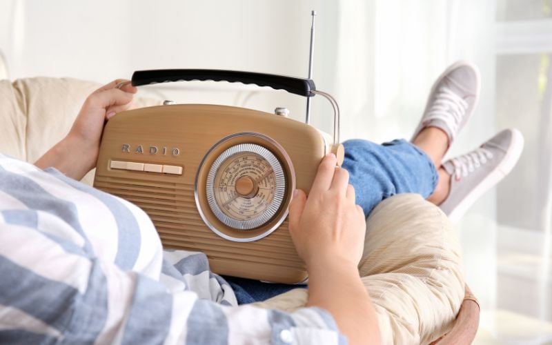 Person sits cross-legged on a couch in a bright room with a portable shortwave radio on his lap